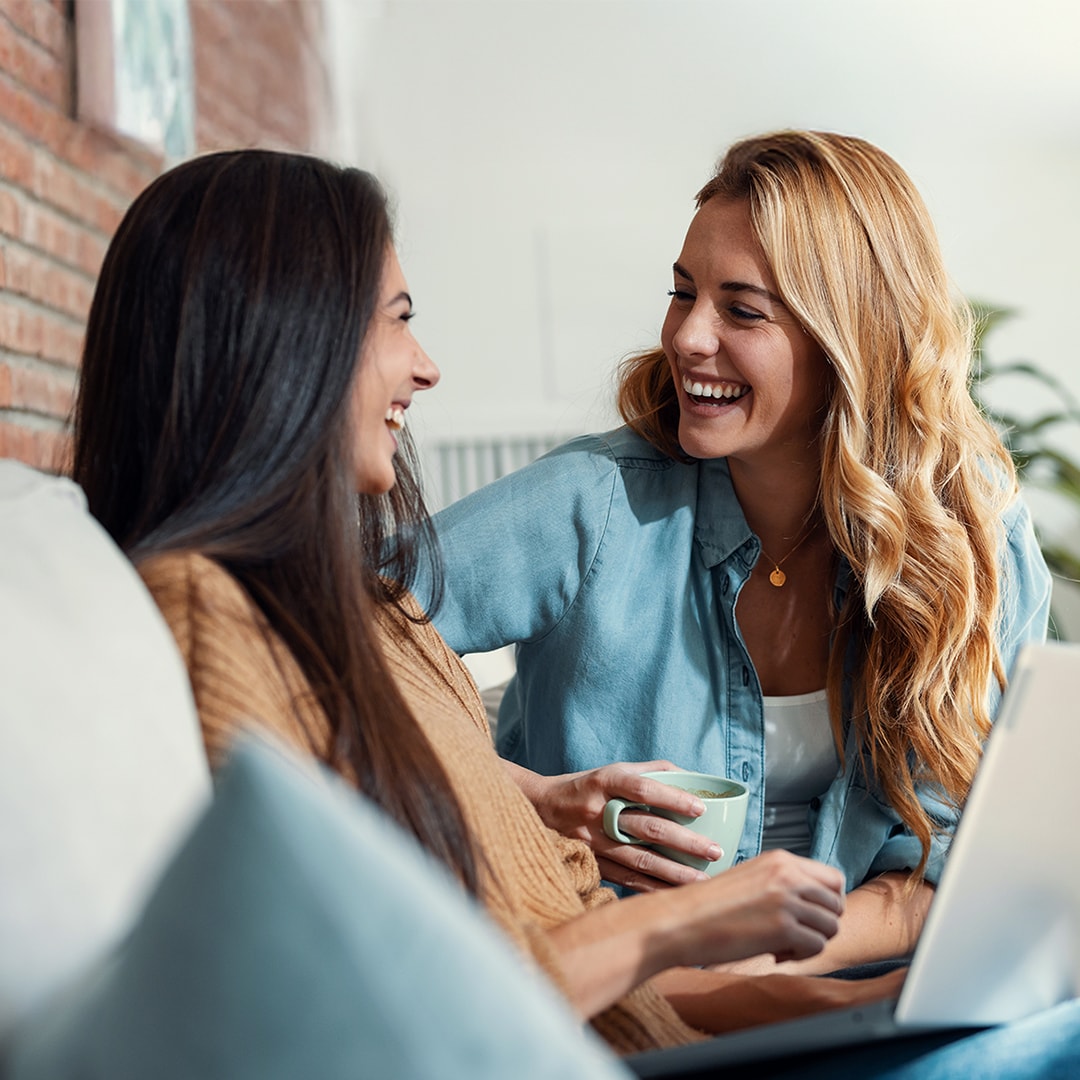 Dos mujeres sonrientes sentadas en un sofá delante de un ordenador portátil 