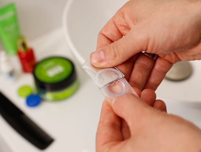 Primer plano de manos abriendo un blíster de lentes de contacto en el baño.