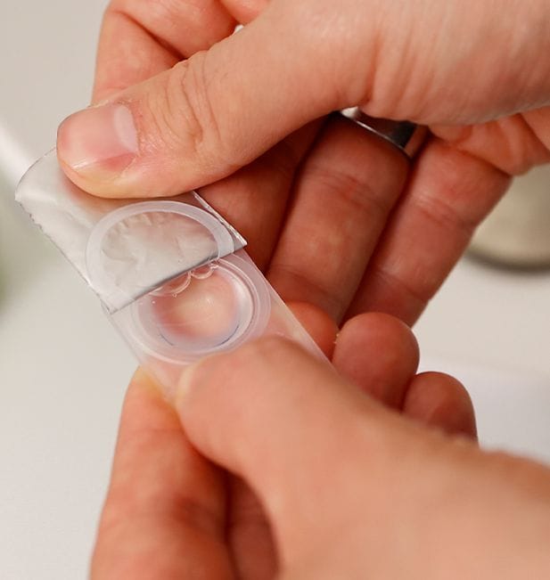 Primer plano de manos abriendo un blíster de lentes de contacto en el baño.